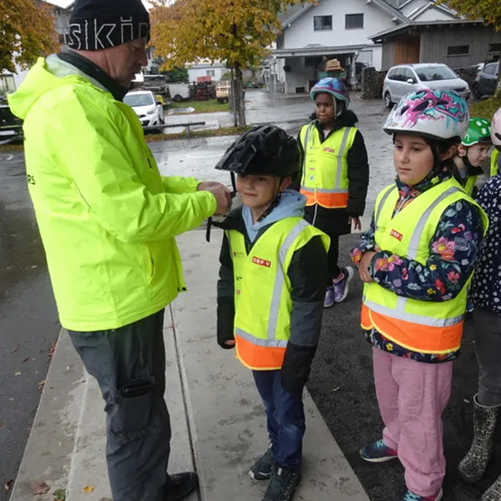 Ein Mann in einer gelben Jacke zeigt Kindern, wie sie ihre Helme aufsetzen. Die Kinder stehen auf dem Bürgersteig, und im Hintergrund befindet sich ein Gebäude.