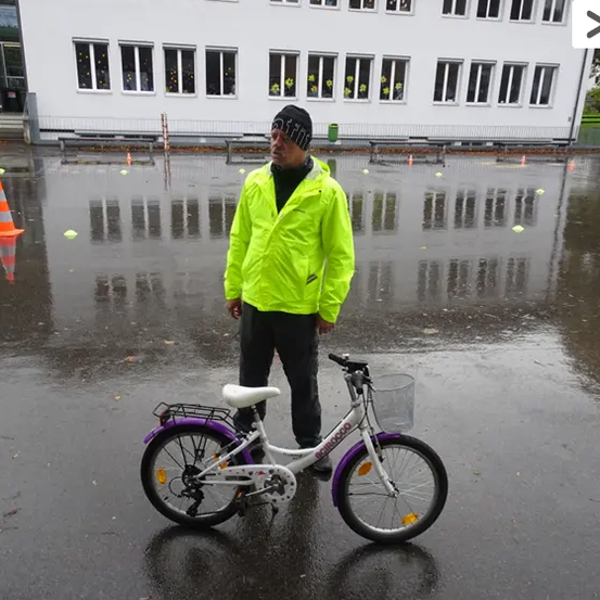 Ein Mann steht mit einem Fahrrad in einem nassen Bereich vor einem Gebäude mit vielen Fenstern. Er trägt eine gelbe Jacke und schwarze Hose.