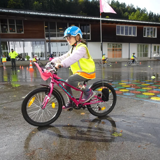 Ein Mädchen in einer neongelben Weste fährt ein pinkes Fahrrad durch einen nassen Bereich mit bemalten Kreisen. Im Hintergrund steht ein Gebäude mit Glasfenstern.