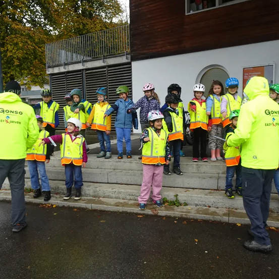 Eine Gruppe von Kindern in gelben Westen, Helmen und Stiefeln steht auf den Stufen mit zwei Erwachsenen.