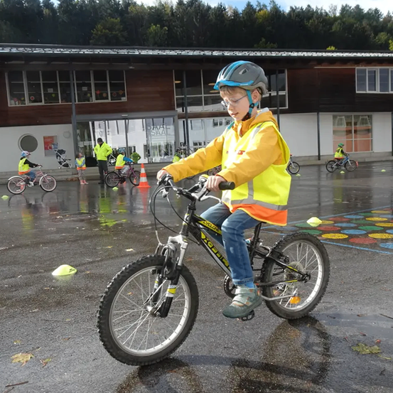 Ein junger Junge fährt auf einer nassen Straße ein Fahrrad, trägt eine Sicherheitsweste und einen Helm. Andere Kinder fahren hinter ihm mit Fahrrädern, mit Verkehrskegeln und einem Gebäude im Hintergrund.