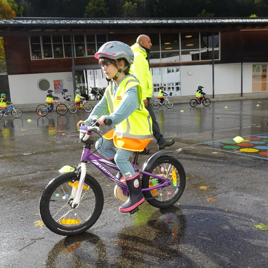 Ein kleines Mädchen fährt ein lila Fahrrad auf einer nassen Straße. Sie trägt eine gelbe Weste und einen Helm. Ein Mann folgt ihr in einem gelben Regenmantel. In der Nähe fahren mehrere Kinder mit Fahrrädern.