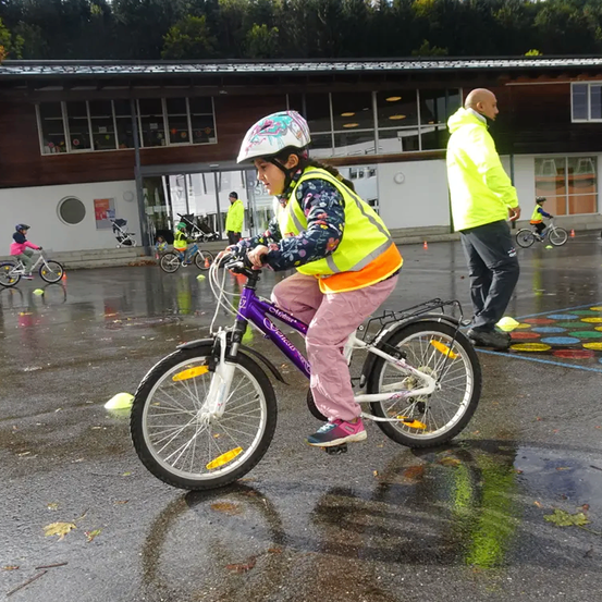 Ein junges Mädchen fährt ein lila Fahrrad auf einer nassen Straße, trägt einen Helm und eine gelbe Weste. Ein Mann in einer gelben Jacke steht in der Nähe, und mehrere andere Kinder fahren im Hintergrund mit ihren Fahrrädern.