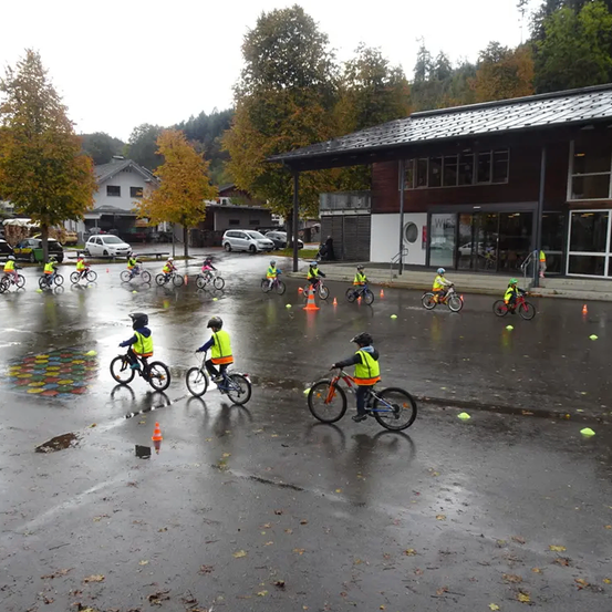 Kinder in Neonwesten üben das Fahrradfahren auf einer nassen Fläche mit Hütchen. Im Hintergrund sind Bäume und geparkte Autos zu sehen.