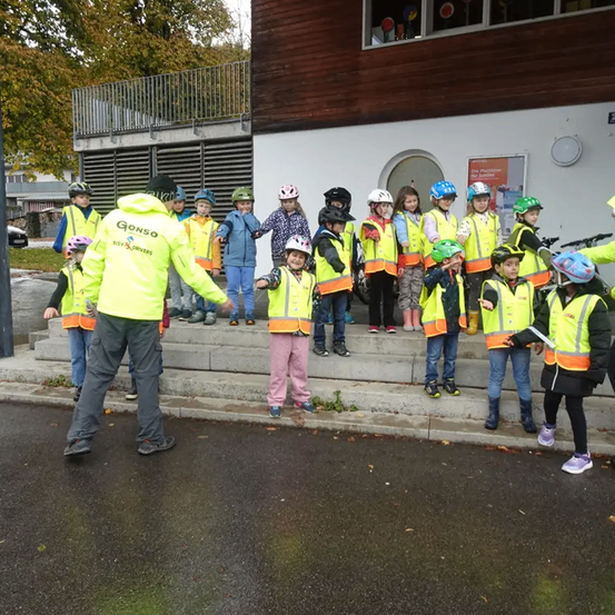 Eine Gruppe von Kindern in Sicherheitswesten und Helmen steht auf Stufen vor einem Gebäude. Ein Mann in einer gelben Jacke steht in der Nähe.