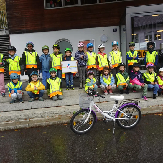 Eine Gruppe von Kindern in Warnwesten und Helmen posiert für ein Foto vor einem Gebäude mit einem Fahrrad im Vordergrund.