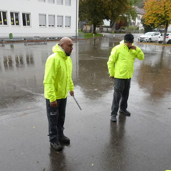 Zwei Männer in neonfarbenen Jacken stehen auf einer nassen Straße, wahrscheinlich nach Regen. Einer hält einen Metallstab. Sie stehen sich gegenüber, wobei Gebäude und Autos im Hintergrund zu sehen sind.