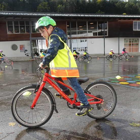 Ein junger Junge fährt ein rotes Fahrrad auf einer nassen Straße, trägt einen Helm und eine reflektierende Weste. Andere Kinder fahren ebenfalls Fahrräder, und Verkehrskegel sind in der Nähe platziert. Im Hintergrund befindet sich ein Gebäude mit Glasfenstern.