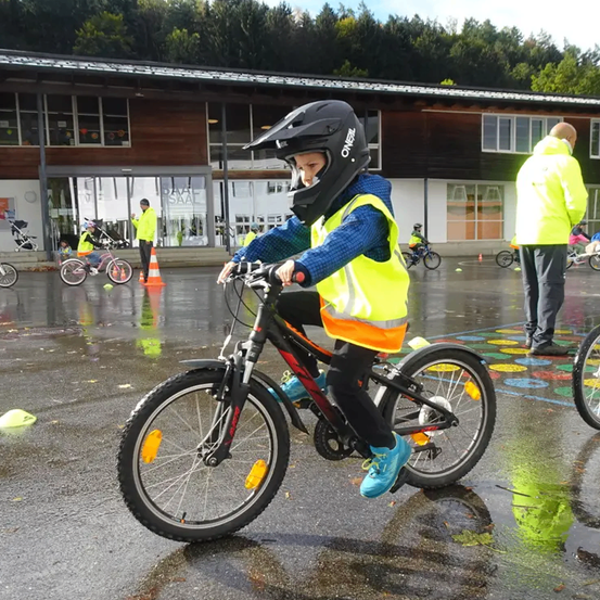 Ein Kind fährt auf einer nassen Straße mit dem Fahrrad, trägt einen Helm und eine Warnweste. Andere Kinder fahren hinter ihm, und ein Mann steht in der Nähe. Im Hintergrund befinden sich Gebäude, Bäume und Verkehrskegel.