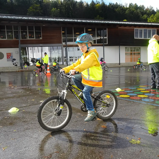 Ein Junge fährt auf einer nassen Straße mit dem Fahrrad. Er trägt eine gelbe Jacke, eine reflektierende Weste und einen Helm. Hinter ihm befindet sich ein Gebäude mit Glasfenstern. Es gibt auch andere Kinder, die Fahrräder fahren. Einige von ihnen tragen Sicherheitswesten. Es gibt auch Verkehrskegel und Blätter auf dem Boden.