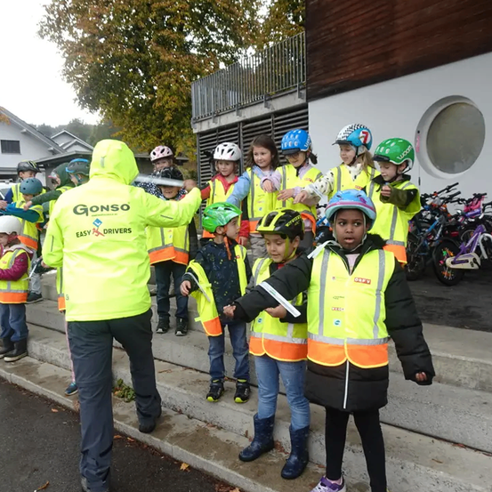 Kinder in neonfarbenen Westen und Helmen versammeln sich auf Treppenstufen mit einer Person in einem Neonwesten, die ein Fahrrad hält. Fahrräder sind hinter ihnen in der Nähe eines Gebäudes geparkt.