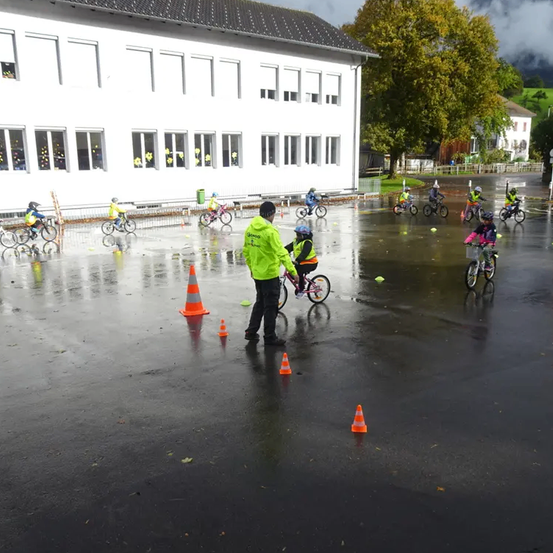 Auf einem regnerischen Parkplatz fahren Kinder mit Helmen Fahrrad. Ein Mann in einer gelben Jacke gibt ihnen Anweisungen. Kegel und Bälle sind verstreut. Ein weißes Gebäude und Bäume sind im Hintergrund.