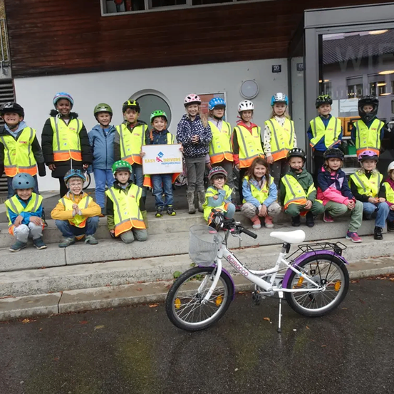 Eine Gruppe von Kindern in gelben Westen posiert mit einem Schild vor einem Gebäude, mit einem Fahrrad auf der Straße.
