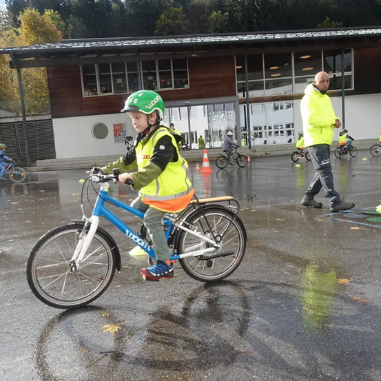 Ein junger Junge fährt auf einer nassen Straße ein blaues Fahrrad, trägt einen grünen Helm und eine gelbe Weste. Ein Erwachsener geht in der Nähe, und mehrere andere Kinder fahren Fahrräder hinter orangefarbenen Verkehrskegeln.