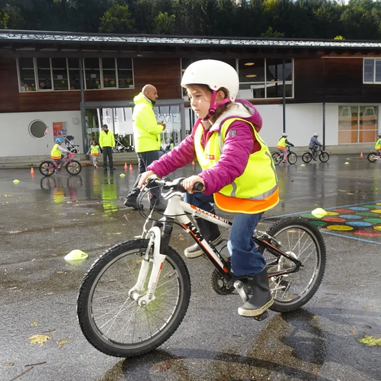 Ein junges Mädchen in einem gelben Weste fährt ein Fahrrad auf einem nassen Gebiet, mit Erwachsenen und anderen Kindern in der Nähe, alle tragen Sicherheitsausrüstung. Gebäude und Bäume sind im Hintergrund.