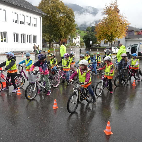 Eine Gruppe von Kindern in Sicherheitswesten und Helmen fährt auf einer nassen Straße mit Fahrrädern, umringt von Verkehrskegeln. Dahinter befinden sich Gebäude und Bäume.