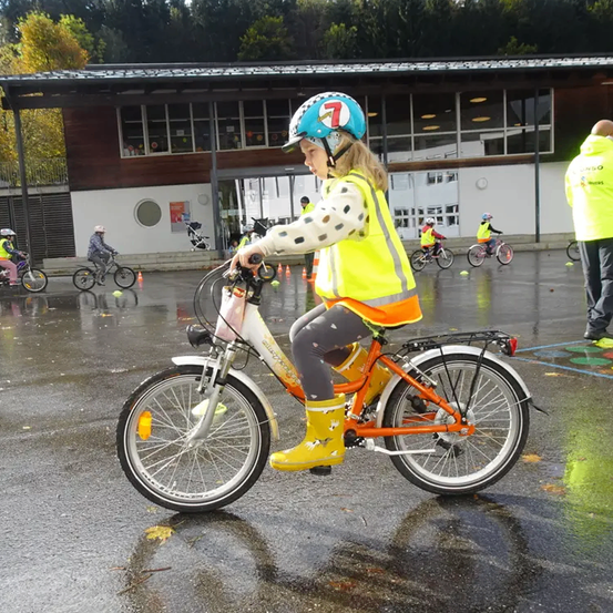 Ein junges Mädchen fährt auf einem Fahrrad in einem Parkplatz und trägt einen Helm und eine reflektierende Weste. Andere Kinder fahren hinter ihr Fahrräder. Der Bereich ist nass, mit Verkehrskegeln und einem Gebäude im Hintergrund.