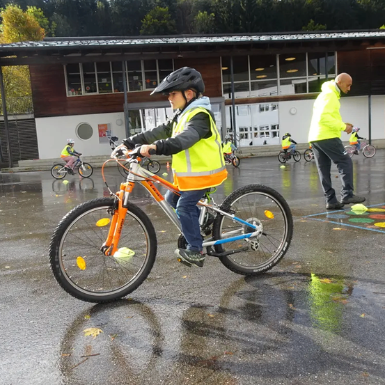 Ein Junge fährt bei Regenwetter mit seinem Fahrrad und trägt eine Sicherheitsweste. In der Nähe steht ein Mann, und andere Kinder fahren ihre Fahrräder. Ein Gebäude mit Glasfenstern ist im Hintergrund.