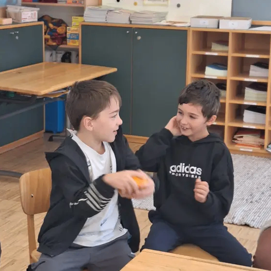 Zwei junge Jungen in einem Klassenzimmer, einer hält eine Orange, lächelt und spielt zusammen. Hinter ihnen stehen Regale mit Büchern und Aufbewahrungsboxen. Eine Tafel zeigt 'Klassenrat', was auf ein Klassenzimmer hinweist.