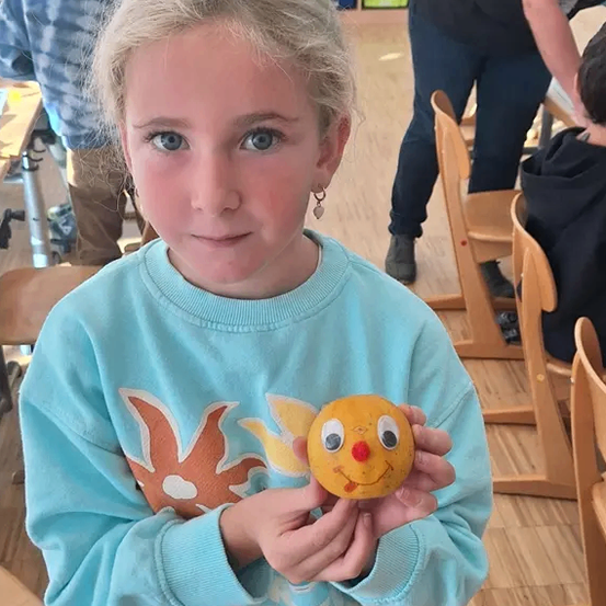 Ein junges Mädchen in einem blauen Shirt hält einen gelben Gegenstand mit einem Gesicht, in einem Klassenzimmer. Hinter ihr steht eine Person und andere sind sitzend.