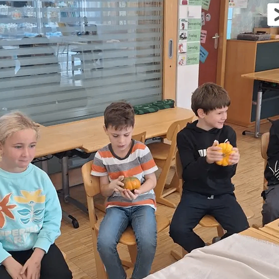 Vier Jungen sitzen auf Stühlen in einem Klassenzimmer und halten kleine Kürbisse. Hinter ihnen sind Tische in Reihen angeordnet. Ein Kalender ist an der Wand angeheftet und in der Nähe befindet sich eine Tafel. Das Klassenzimmer hat Holzböden und Glasfenster.