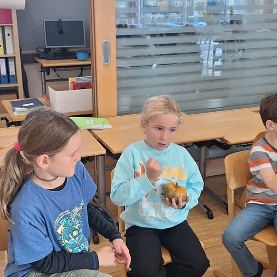 Drei Kinder sitzen in einem Klassenzimmer. Eines hält eine Kürbis und redet. Ein anderes schaut zu, während das dritte zuhört. Hinter ihnen sind Schreibtische und ein Fenster.