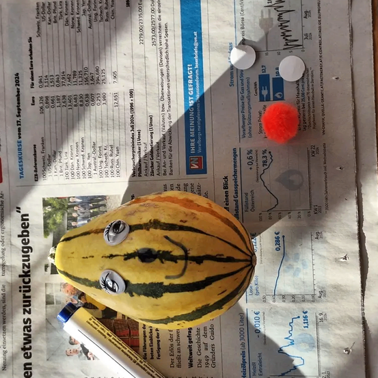 Ein bemalter Kürbis mit Augen und Mund sitzt auf einer Zeitung mit einem Diagramm, zwei weißen Knöpfen und einem roten Pompon.