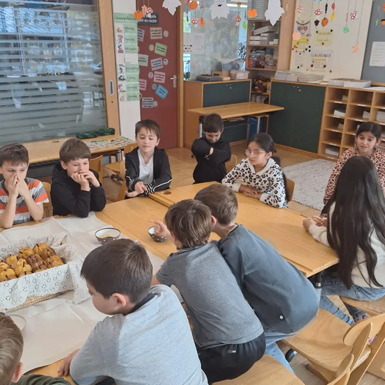 Eine Gruppe von Kindern sitzt in einem Klassenzimmer um einen Tisch und isst. Ein Korb mit Gebäck steht auf dem Tisch. Sie sind lässig gekleidet. Der Raum ist mit verschiedenen Gegenständen dekoriert.