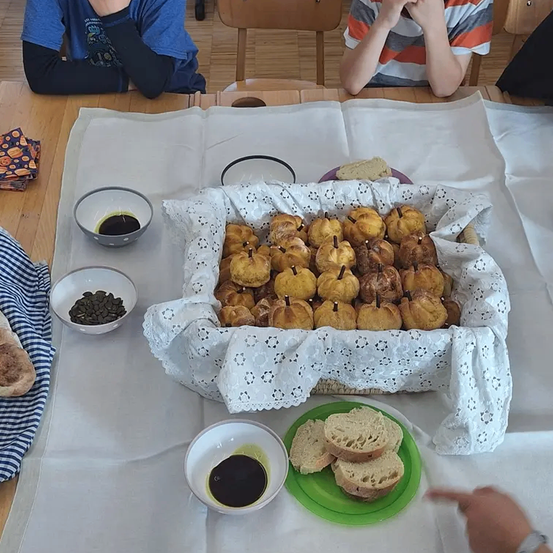Ein Tisch mit einem weißen Tuch zeigt einen Korb mit goldbraunen Gebäckstücken, eine Tasse Kaffee und eine Schale mit Bohnen. Zwei Kinder sitzen in der Nähe, und eines hält ein Sandwich.