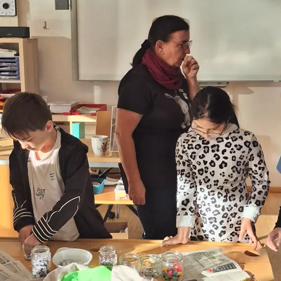 Eine Frau und zwei Kinder sind in einem Klassenzimmer. Die Frau steht hinter einem Schreibtisch, und die Kinder stehen vor einem Tisch mit verschiedenen Gegenständen.