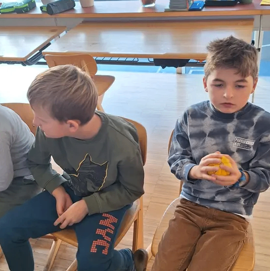 Zwei Jungen sitzen auf Stühlen in einem Klassenzimmer. Ein Junge hält eine Orange, während der andere Junge nach unten schaut.