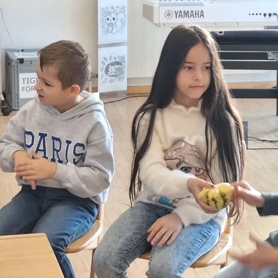 Zwei Kinder sitzen auf Stühlen. Ein Mädchen hält eine Kürbis und ein Junge schaut sie an. Dahinter ist ein Yamaha-Klavier und Poster zu sehen.