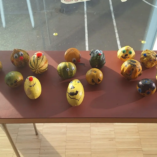 Ein Tisch zeigt eine Sammlung geschnitzter Kürbisse und Gourden, jeder mit verspielten Gesichtern bemalt, in der Nähe eines Fensters, das einen Parkplatz zeigt.