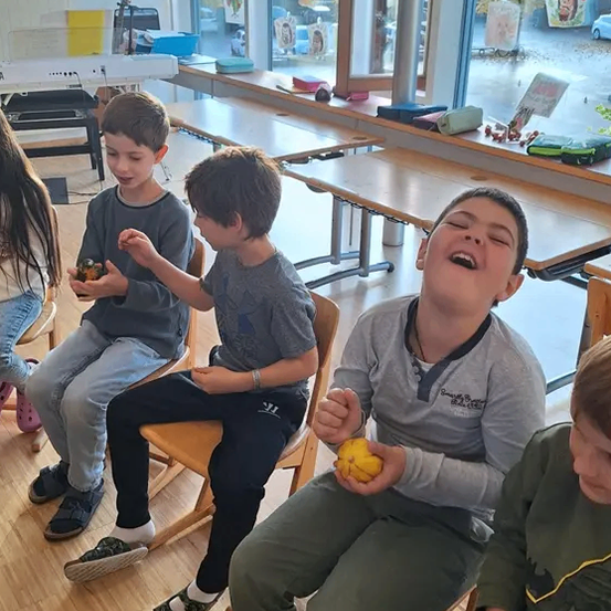 Vier Jungen sitzen in einem Klassenzimmer, einer hält ein kleines gelbes Objekt, während ein anderer Junge lacht. Hinter ihnen sind Tische und Stühle angeordnet. Ein Fenster zeigt einen Blick auf einen regnerischen Tag draußen.
