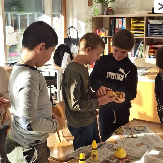 Drei Jungen stehen um einen Tisch mit gelben Gegenständen und Zeitungen. Sie scheinen zu malen, einer hält ein gelbes Objekt. Ein Regal und ein Kreuz sind im Hintergrund.