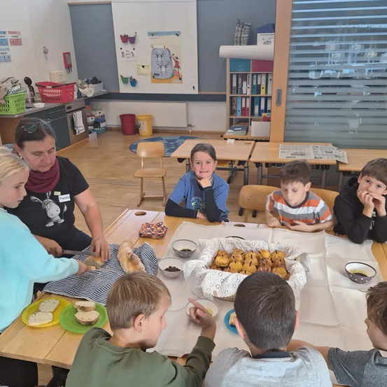 Eine Gruppe von Kindern und einer Frau sitzen um einen Tisch in einem Klassenzimmer. Sie essen Brot und es gibt Schüsseln mit Essen auf dem Tisch.