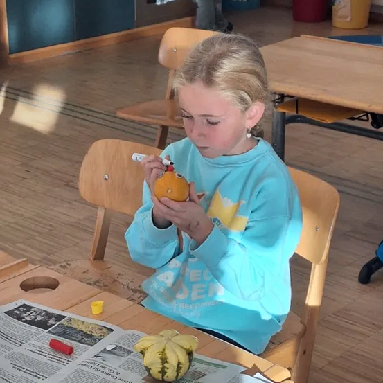 Ein Mädchen sitzt an einem Tisch und malt eine Kürbis. Sie trägt ein blaues Sweatshirt. Hinter ihr steht ein Holztisch und ein Stuhl, und in der Ferne steht eine Person.