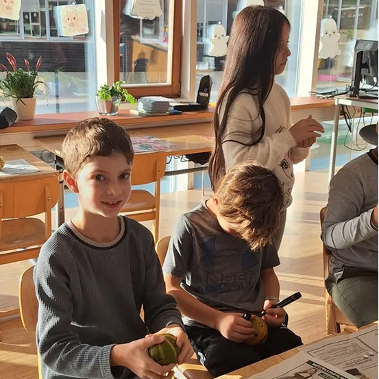 Zwei Jungen sitzen an einem Tisch, einer hält ein grünes Objekt. Ein Mädchen steht hinter ihnen, und ein weiteres Mädchen sitzt rechts. Der Raum hat einen Tisch, Stühle, einen Monitor, eine Topfpflanze und Fenster mit Dekorationen.