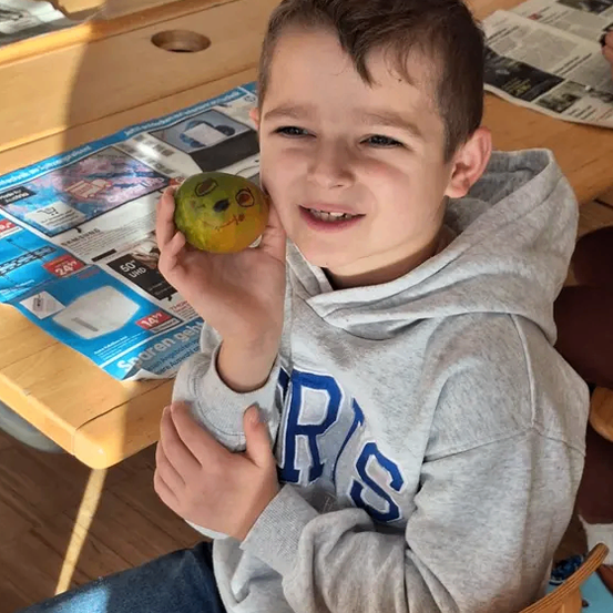 Ein Junge in einer grauen Kapuzenjacke sitzt an einem Schreibtisch und hält einen grünen Apfel mit einem gezeichneten Gesicht.