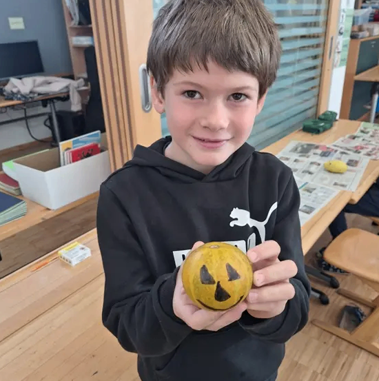 Ein junger Junge in einer schwarzen Kapuzenjacke hält eine geschnitzte Kürbis in einem Klassenzimmer.