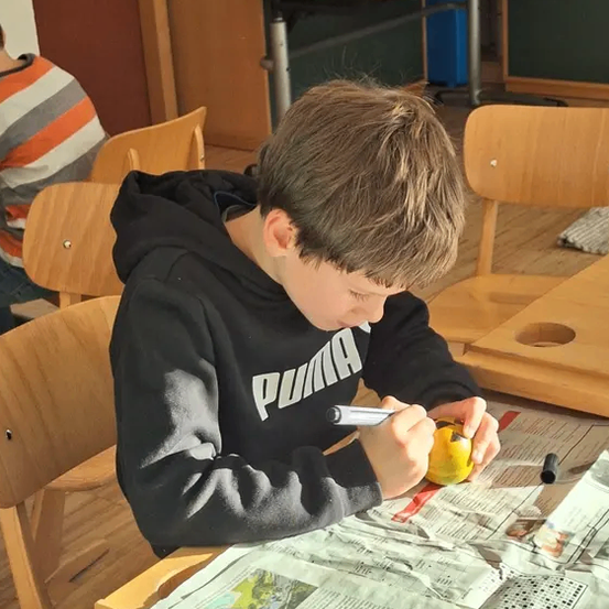 Ein Junge in einem schwarzen Puma-Hoodie schneidet mit einem Messer eine Zitrone auf einer Zeitung aus. Er sitzt an einem Tisch mit Holzstühlen.