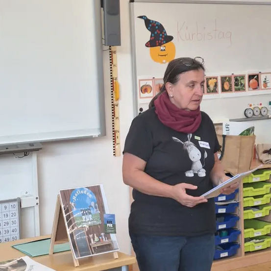 Eine erwachsene Frau steht vor einem Klassenzimmer, hält ein Papier in der Hand und liest. Hinter ihr befindet sich eine Tafel mit einer Kürbiszeichnung. Ein Buch und ein Schreibtisch sind im Vordergrund.