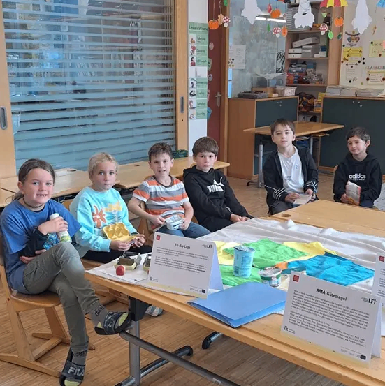 Eine Gruppe von Kindern sitzt in einem Klassenzimmer um einen Tisch herum und beschäftigt sich mit einem Projekt. Verschiedene Materialien liegen auf dem Tisch, und ein Mädchen hält eine Flasche. Hinter ihnen führt eine Glastür in einen Flur.