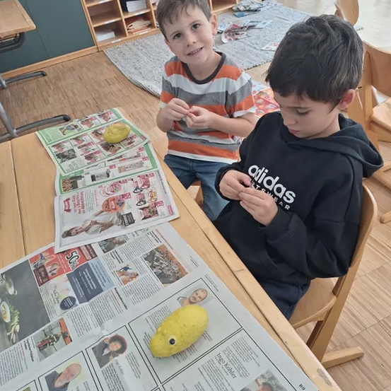 Zwei Jungen sitzen an einem Tisch mit Zeitungen, einer hält ein gelbes Objekt, wahrscheinlich ein Spielzeug, in einem Klassenzimmer mit Holzmöbeln und einem Teppich.
