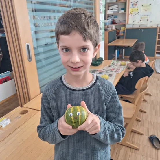 Ein junger Junge in einem blauen Pullover hält eine dekorierte Kürbis mit Augen. Hinter ihm sitzen andere Kinder in einem Klassenzimmer an Tischen. Der Raum ist mit verschiedenen Dekorationen gefüllt.