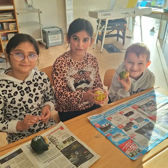 Drei Kinder sitzen an einem Tisch mit einer Zeitung, eines hält eine Kürbis, ein anderes hält einen grünen Apfel und das dritte einen Apfel. Sie sind in einem Raum mit einem Regal, einem Klavier und einer Tafel.