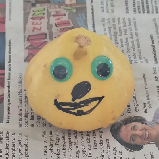 Eine Kartoffel mit einem darauf gezeichneten Gesicht, mit grünen Augen und einem Lächeln, liegt auf einer Zeitung. Die Kartoffel ist gelb und ruht auf einer Zeitung mit deutschem Text.