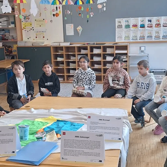Eine Gruppe von Kindern in einem Klassenzimmer sitzt um einen Tisch herum und ist möglicherweise an einem Projekt beteiligt. Papiere mit dem Text 'AMA Biosiegel' liegen auf dem Tisch. Es gibt farbenfrohe Dekorationen an der Wand.