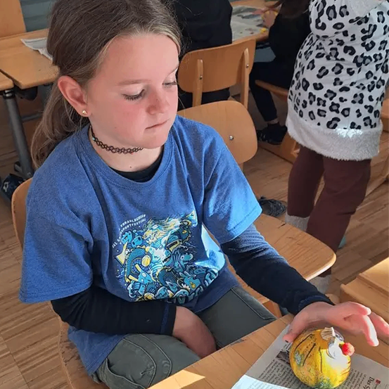 Ein junges Mädchen sitzt an einem Schreibtisch, die Augen geschlossen, und hält ein gelbes Ei. Hinter ihr sitzt ein anderer Schüler an einem Tisch, und eine Person in einem Leopardenmuster-Kleid steht in der Nähe.