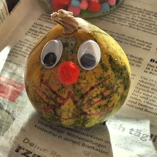 Eine Kürbis mit bemaltem Gesicht, mit zwei Augen und einer roten Nase, liegt auf einer Zeitung mit Text. Dahinter steht ein Glas mit bunten Kugeln auf einer Holzoberfläche.
